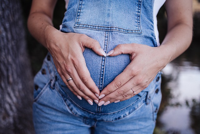 Pregnant woman dressed in blue denim overalls holding her stomach with her hands in the shape of a heart 